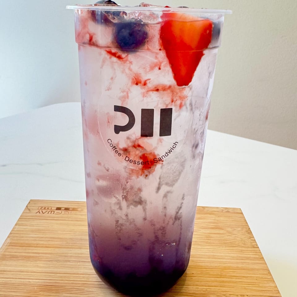 A clear plastic cup of berry soda with ice, blueberries, and a strawberry, on a wooden board.