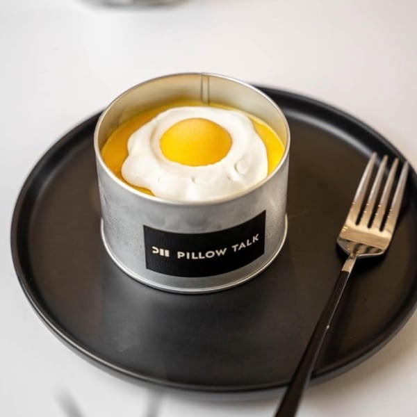Pillow Talk Cafe's egg-shaped dessert in a metal tin, resembling a fried egg, on a black plate with a fork.