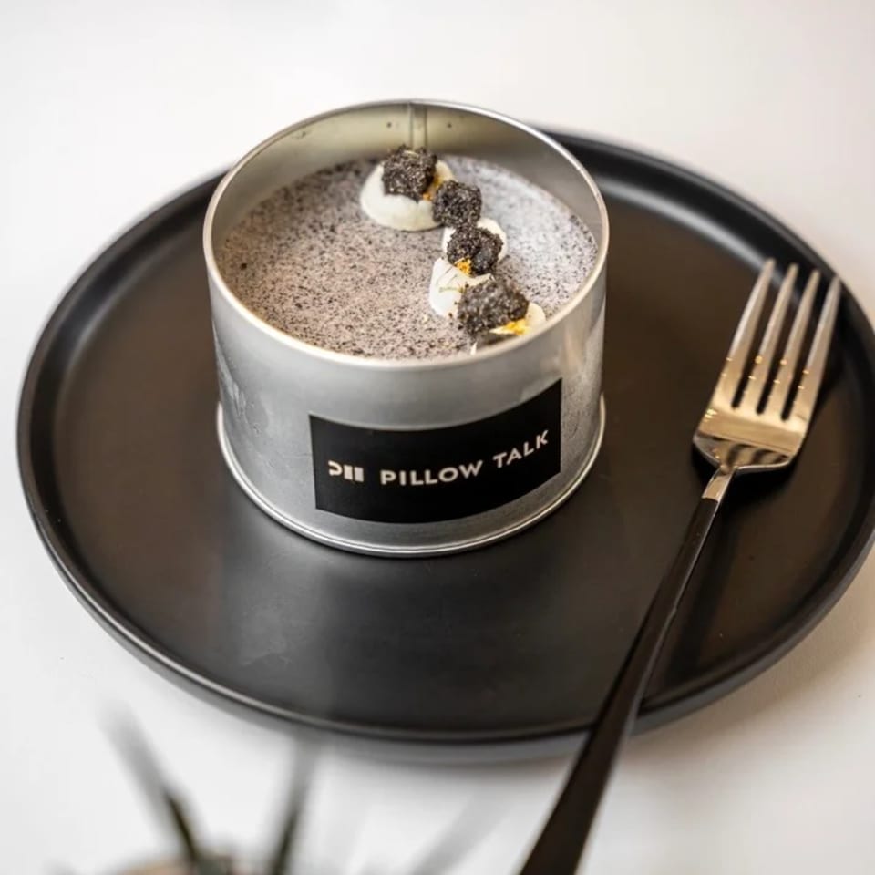 Pillow Talk Cafe's dessert in a metal tin, topped with white cream and dark crumbles, on a black plate with a fork.